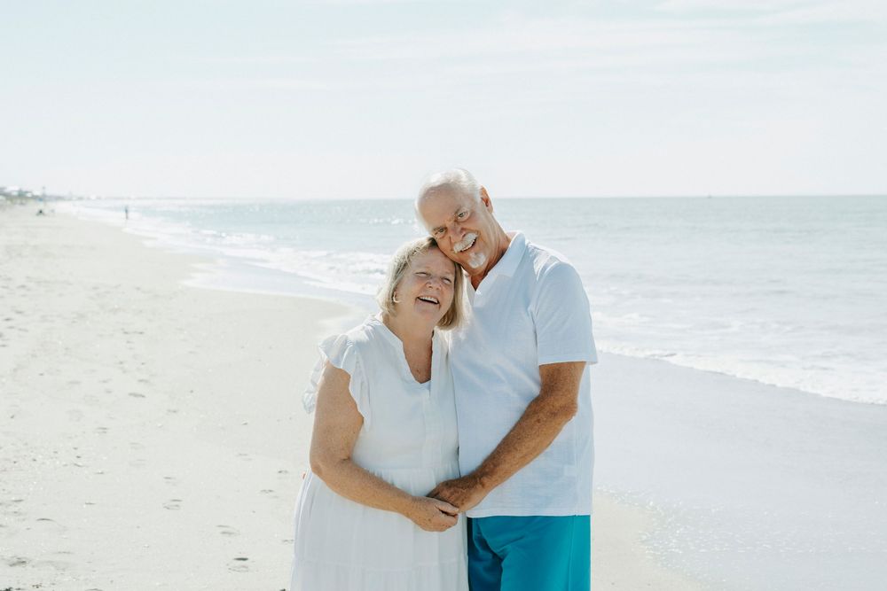 Happy senior couple embracing and smiling on a sunny beach, wearing white and turquoise outfits with ocean waves behind them.