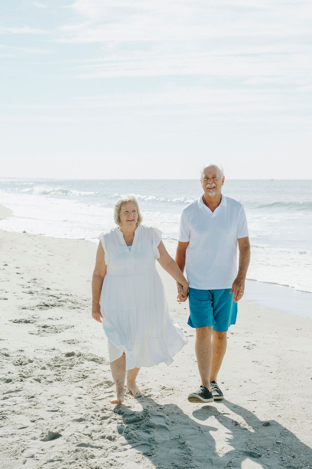 Happy senior couple holding hands and walking barefoot on a sunny sandy beach with ocean waves in the background.