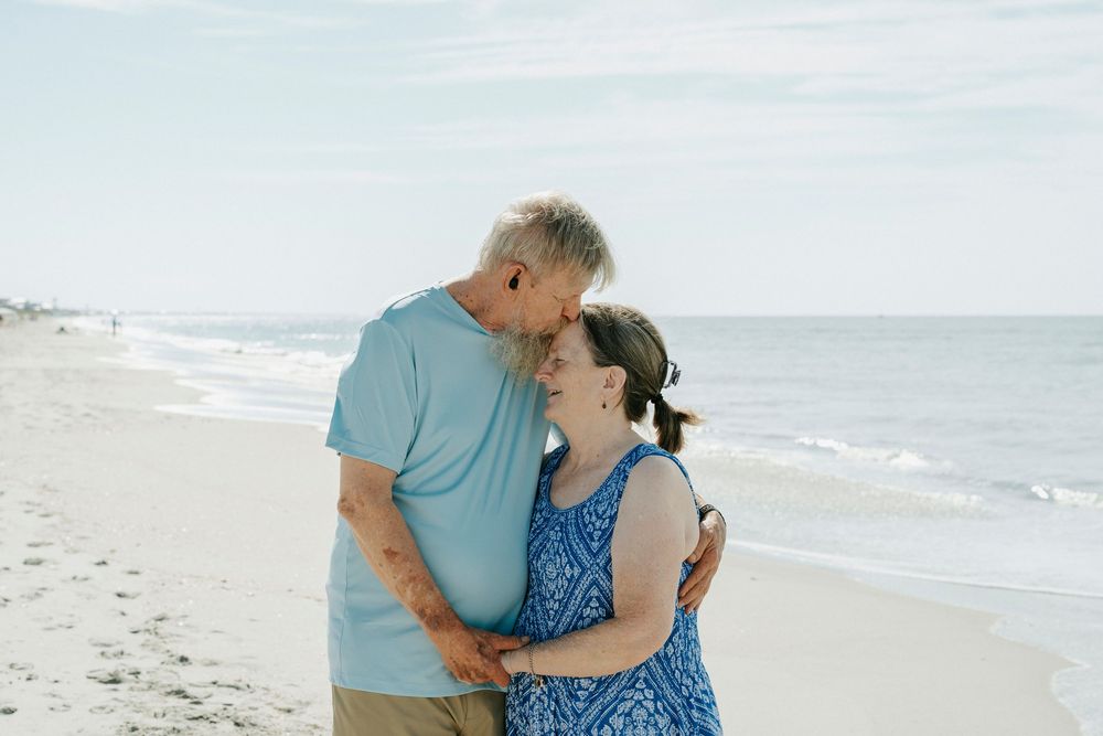Older couple sharing a romantic moment on a sandy beach, embracing and smiling with ocean waves in the background.