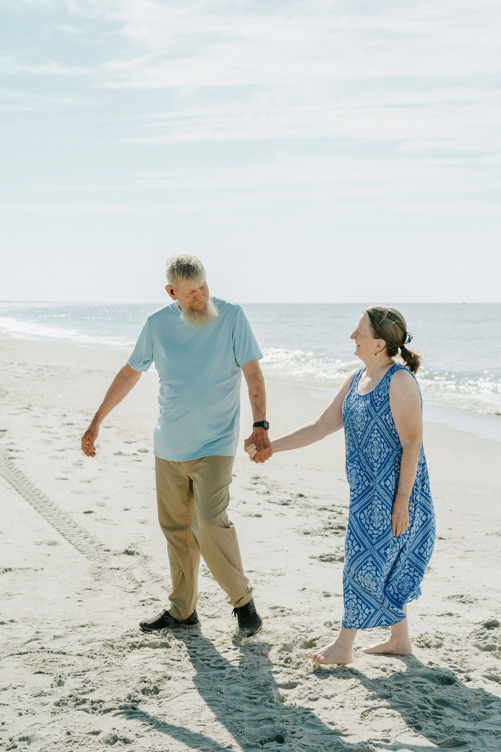 Elderly couple holding hands and walking barefoot on a sunny beach, wearing casual blue outfits with ocean waves behind them.