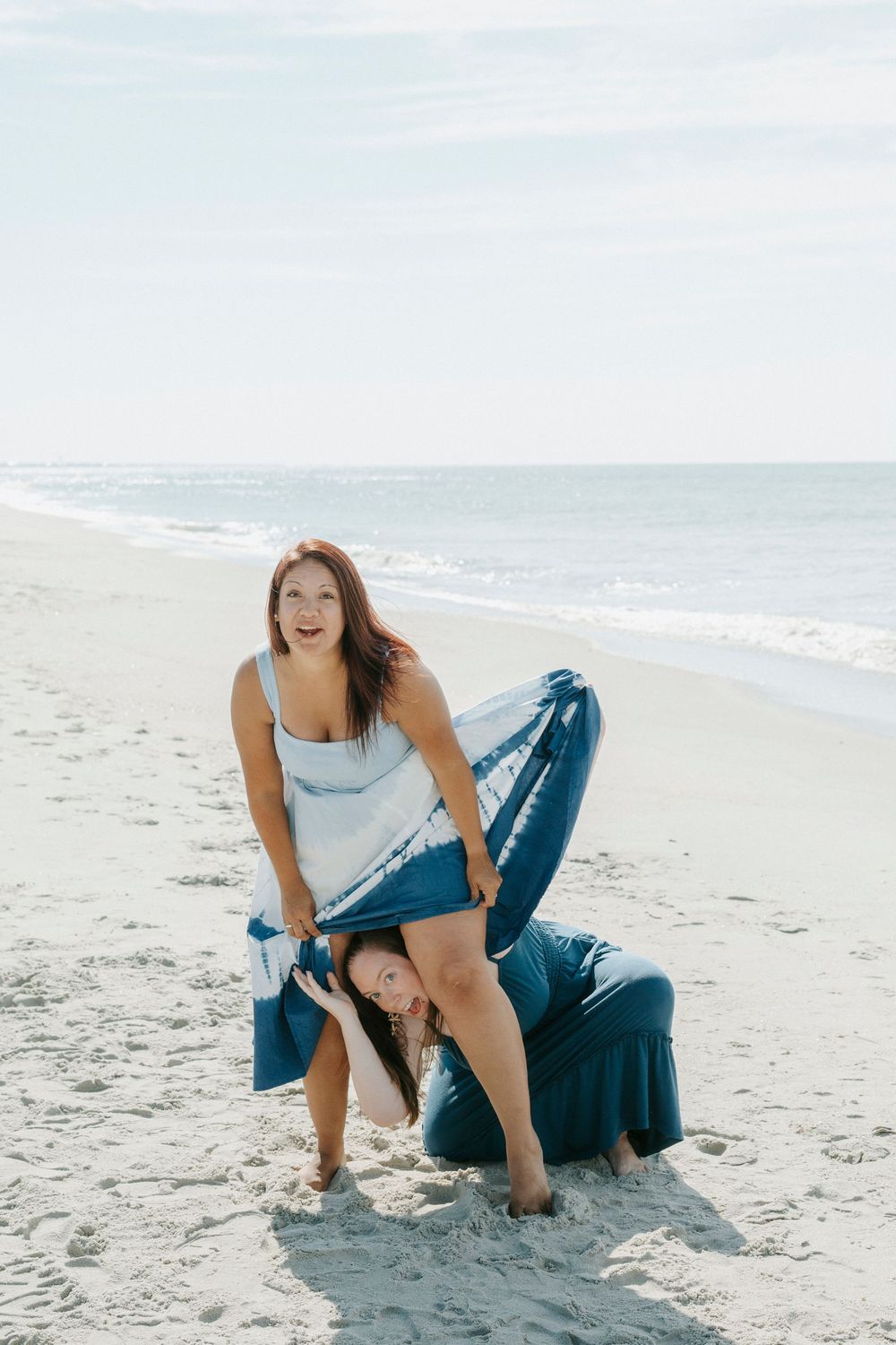 Two women in blue tie-dye outfits playfully posing on a sandy beach with ocean waves in the background.