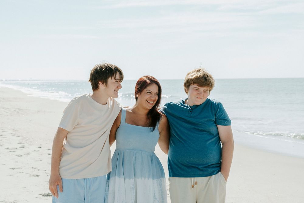 A woman and two teenage boys smile and walk together on a sunny beach with calm ocean waves in the background.