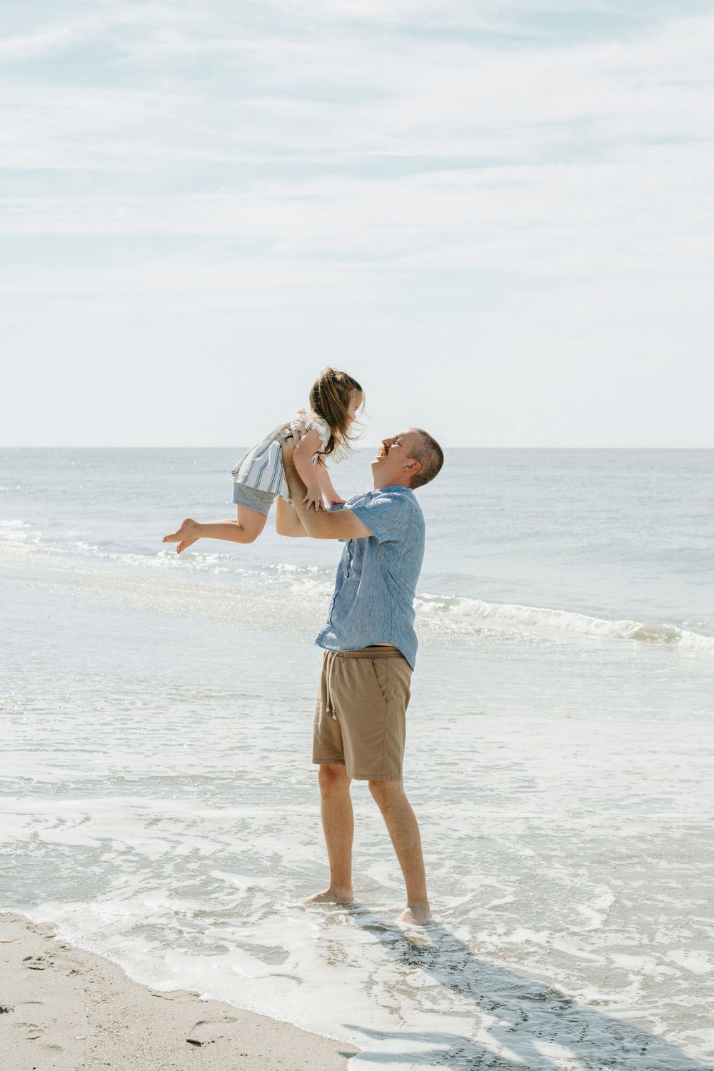 A father joyfully lifts his young child in the air while standing on a sunny beach with ocean waves behind them.