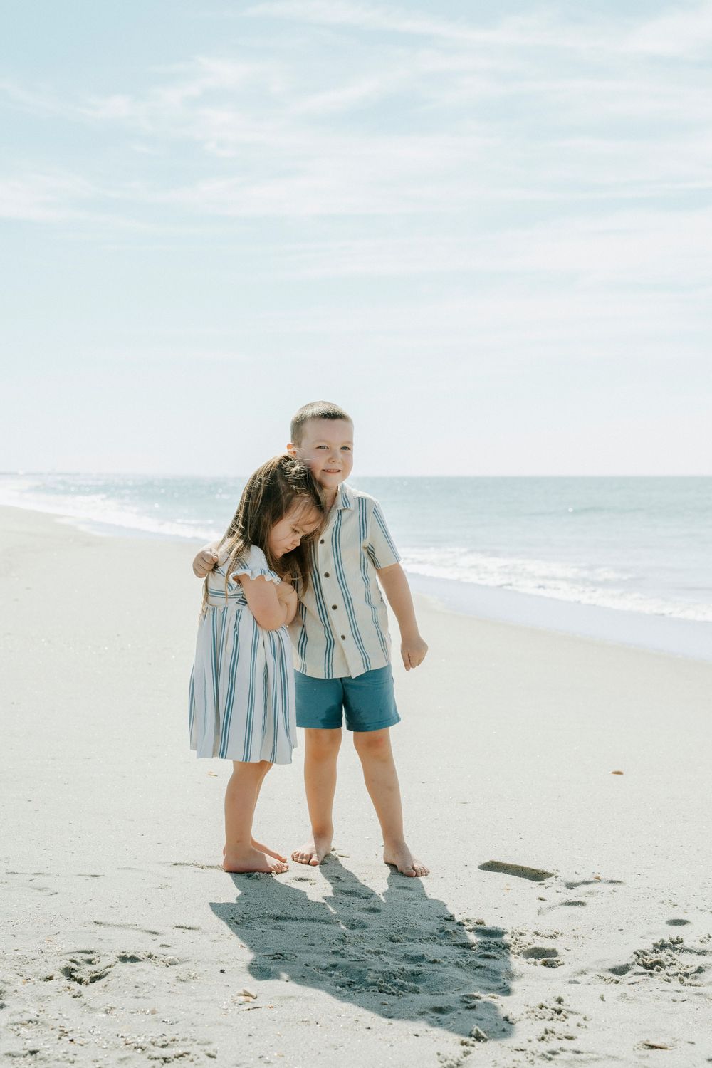 Two children in matching blue striped outfits hugging and smiling on a sunny beach with ocean waves behind them.