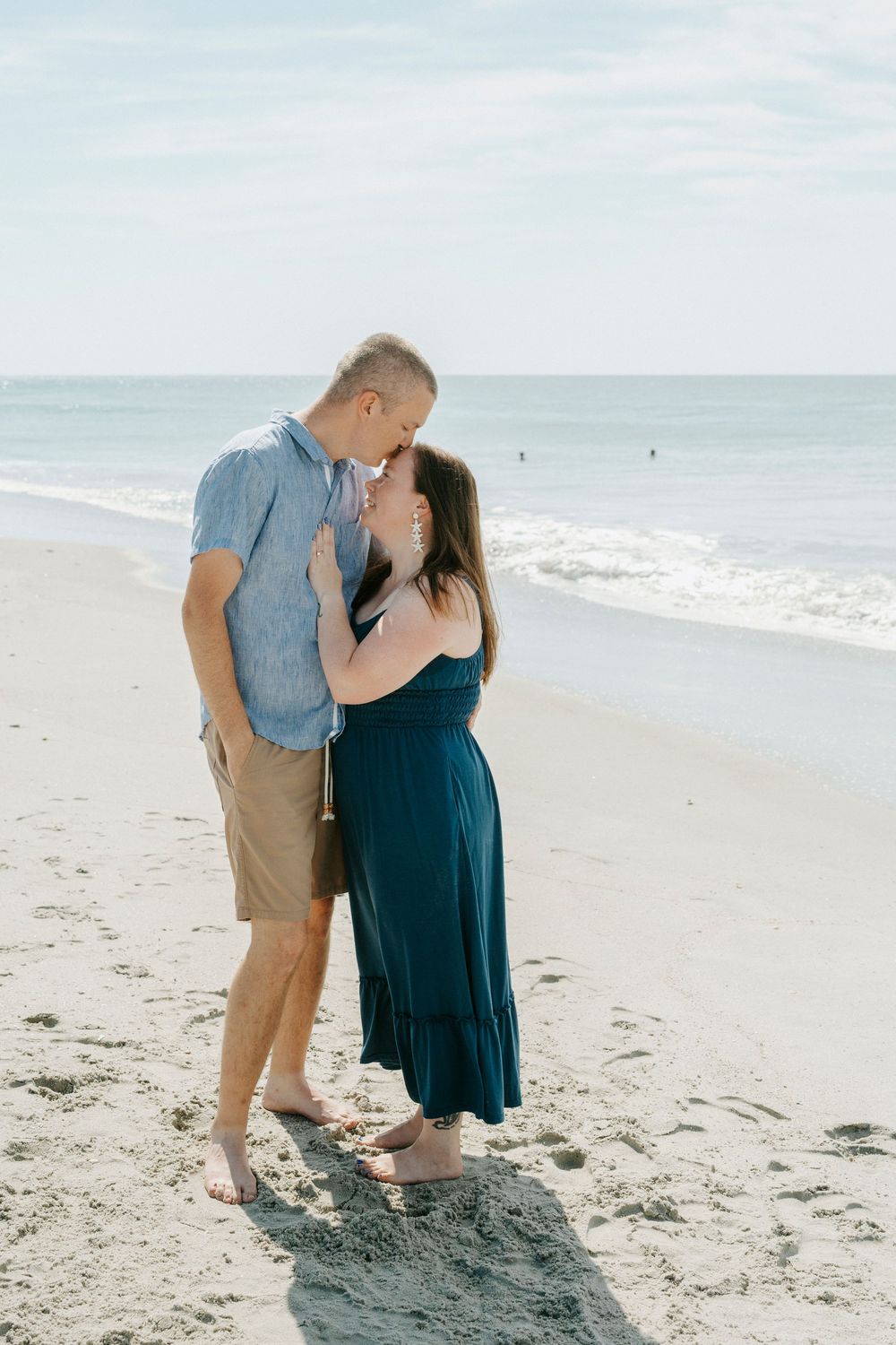 Couple sharing a romantic moment on a sandy beach, woman in teal dress and man in blue shirt, ocean waves behind them.