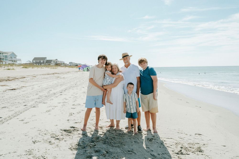 Family of six posing together on a sunny beach with ocean waves and coastal homes in the background.