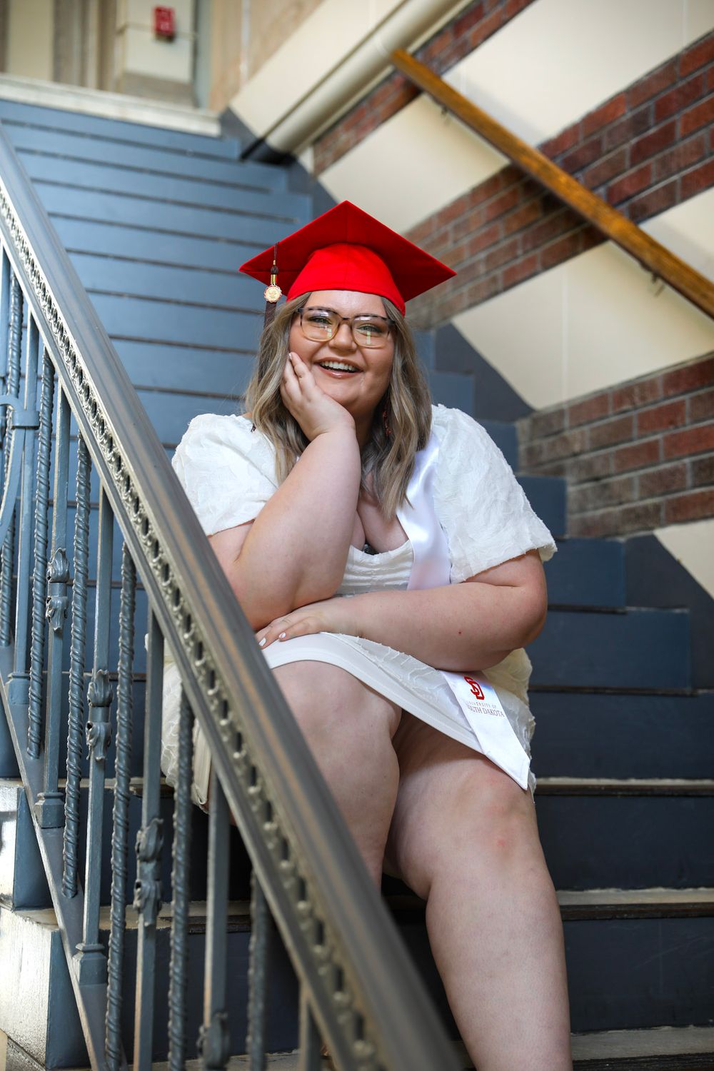 Smiling graduate in red cap and white dress sitting on stairs, celebrating academic achievement.