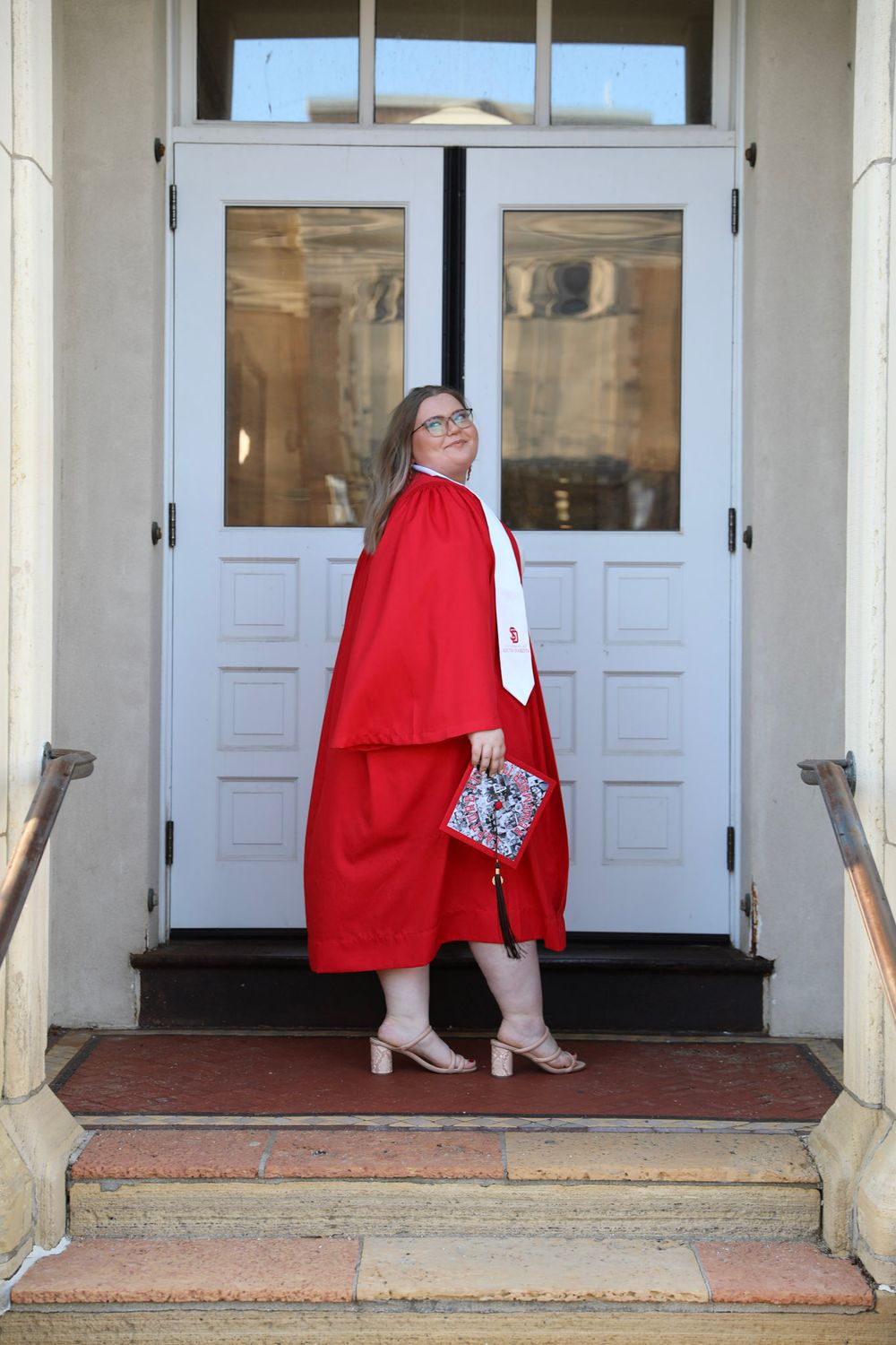 A young woman in a red graduation gown and cap stands on steps before white doors, holding a decorated cap.