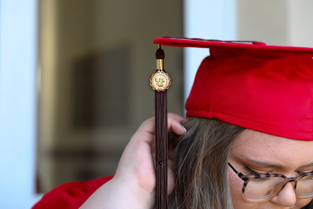 Close-up of a red graduation cap with gold medallion tassel being adjusted on a graduate's head.