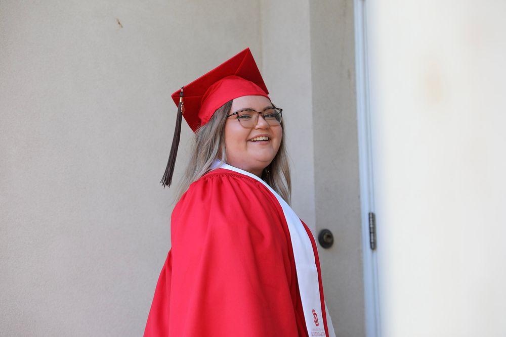 Smiling graduate in red cap and gown posing by a door on graduation day.