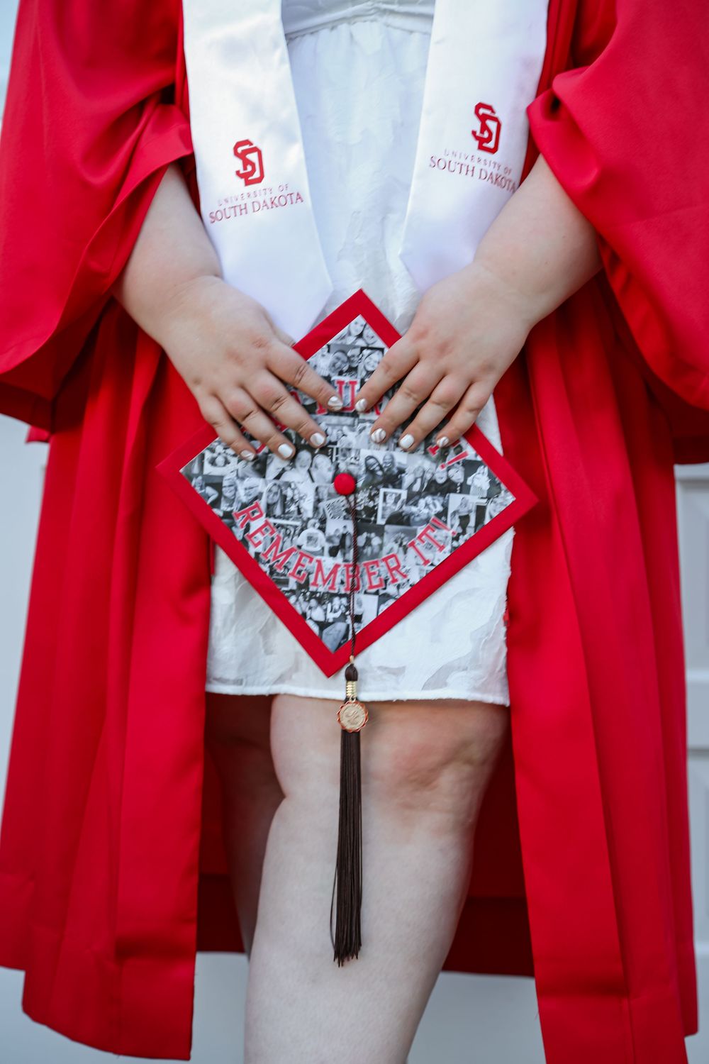University of South Dakota graduate in red robe holds decorated mortarboard cap with photos and tassel.