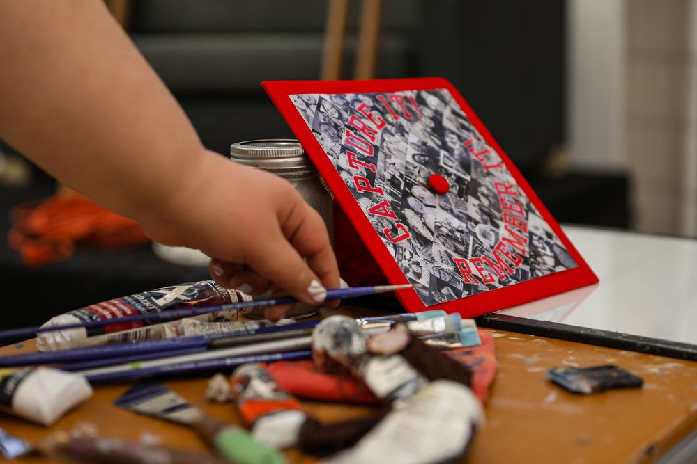 Hand holding colorful abstract red-framed artwork on a cluttered art studio table with brushes and supplies.