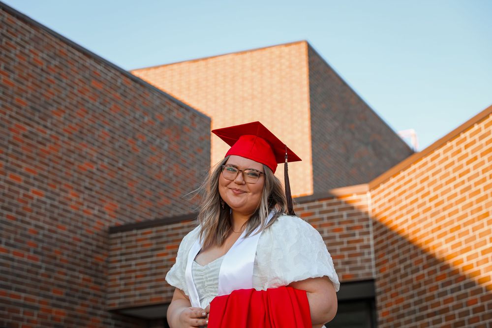 A smiling graduate in red cap and gown holds diploma outside brick building at sunset.