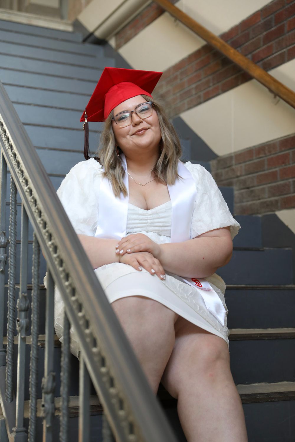Young woman in red graduation cap and white dress sitting on stairs, smiling and celebrating her achievement.