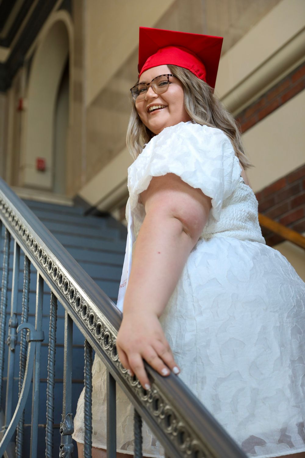 Smiling graduate in red cap and white dress poses on staircase railing during graduation photo session.