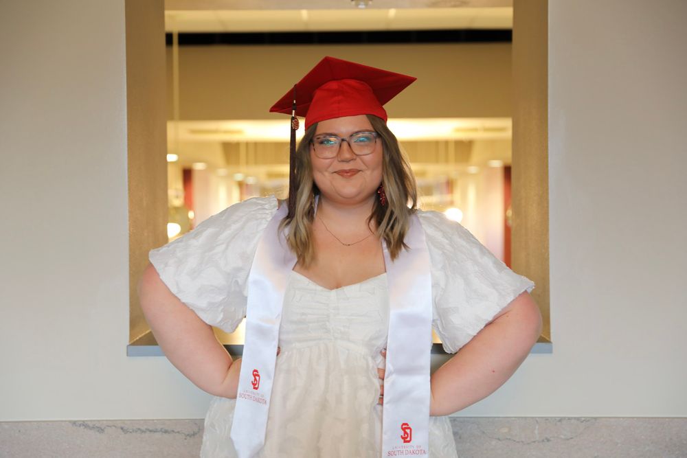 Young woman in red graduation cap and white dress with honor stole smiles proudly at graduation ceremony.