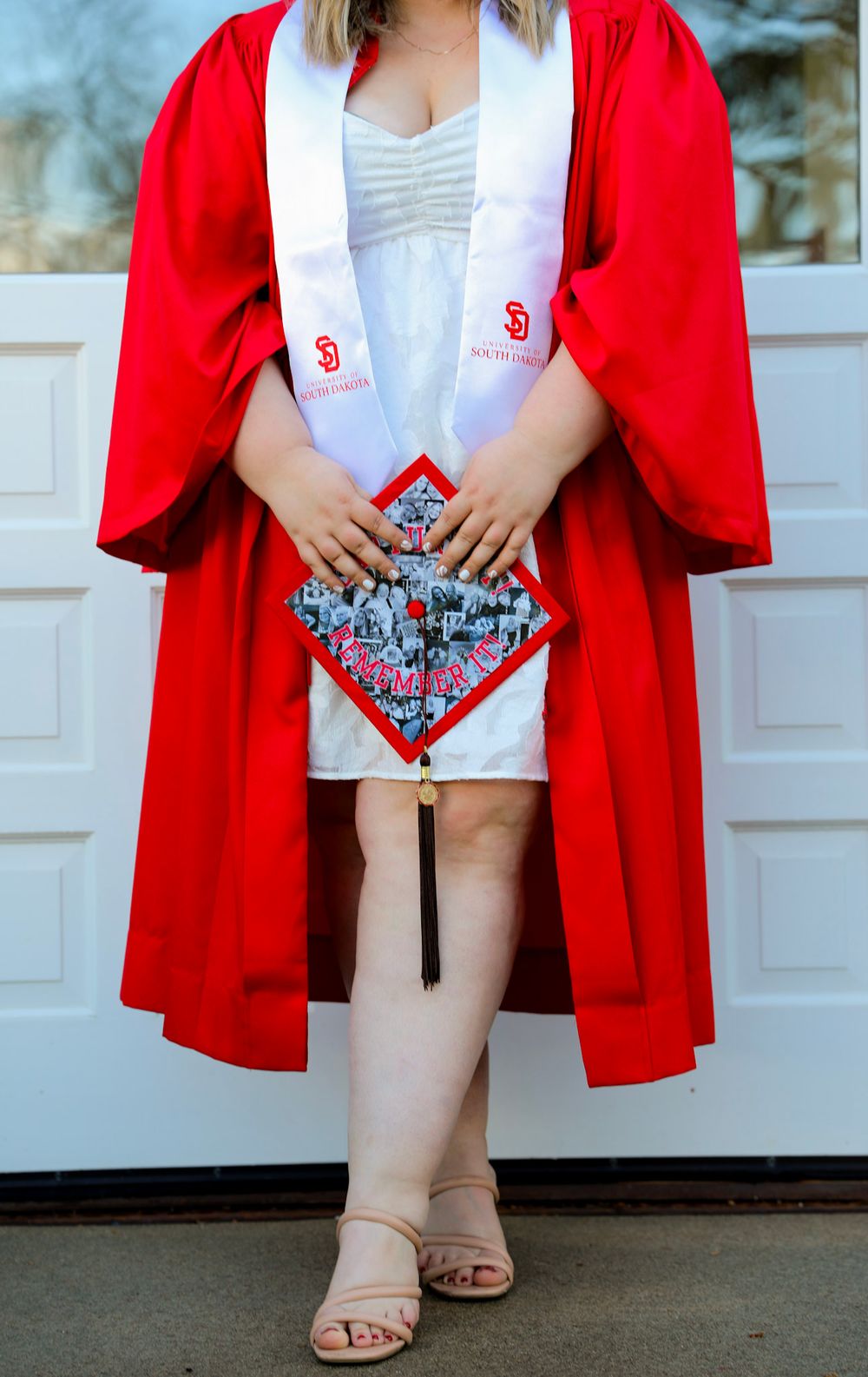 Woman in red graduation gown and white stole holding decorated cap, standing in front of garage door.
