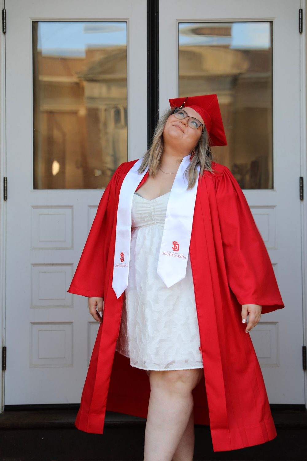 Young woman in red graduation cap and gown with white stole poses confidently before white double doors.
