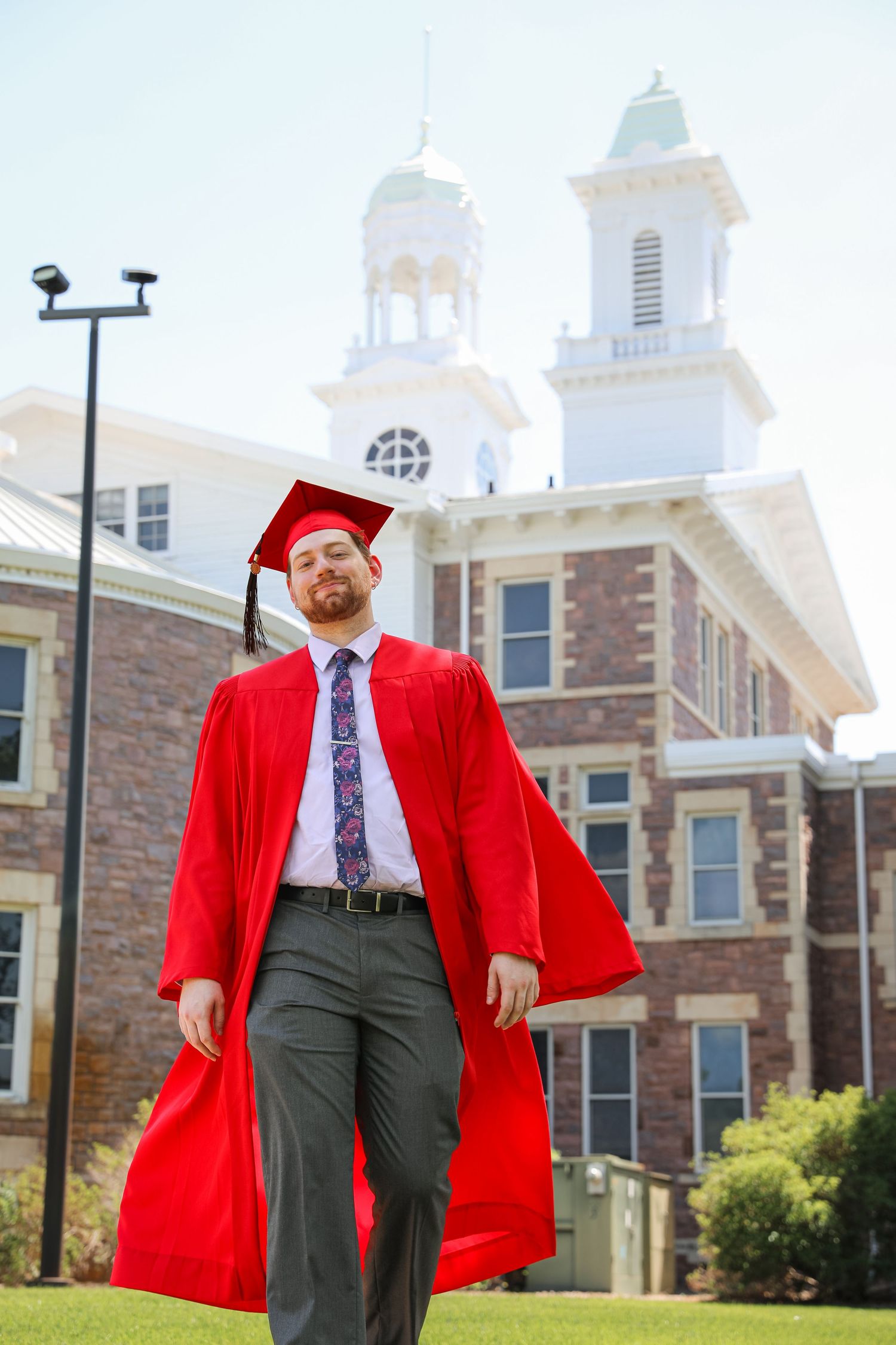 A proud male graduate walks in a red cap and gown in front of a historic brick university building on a sunny day.