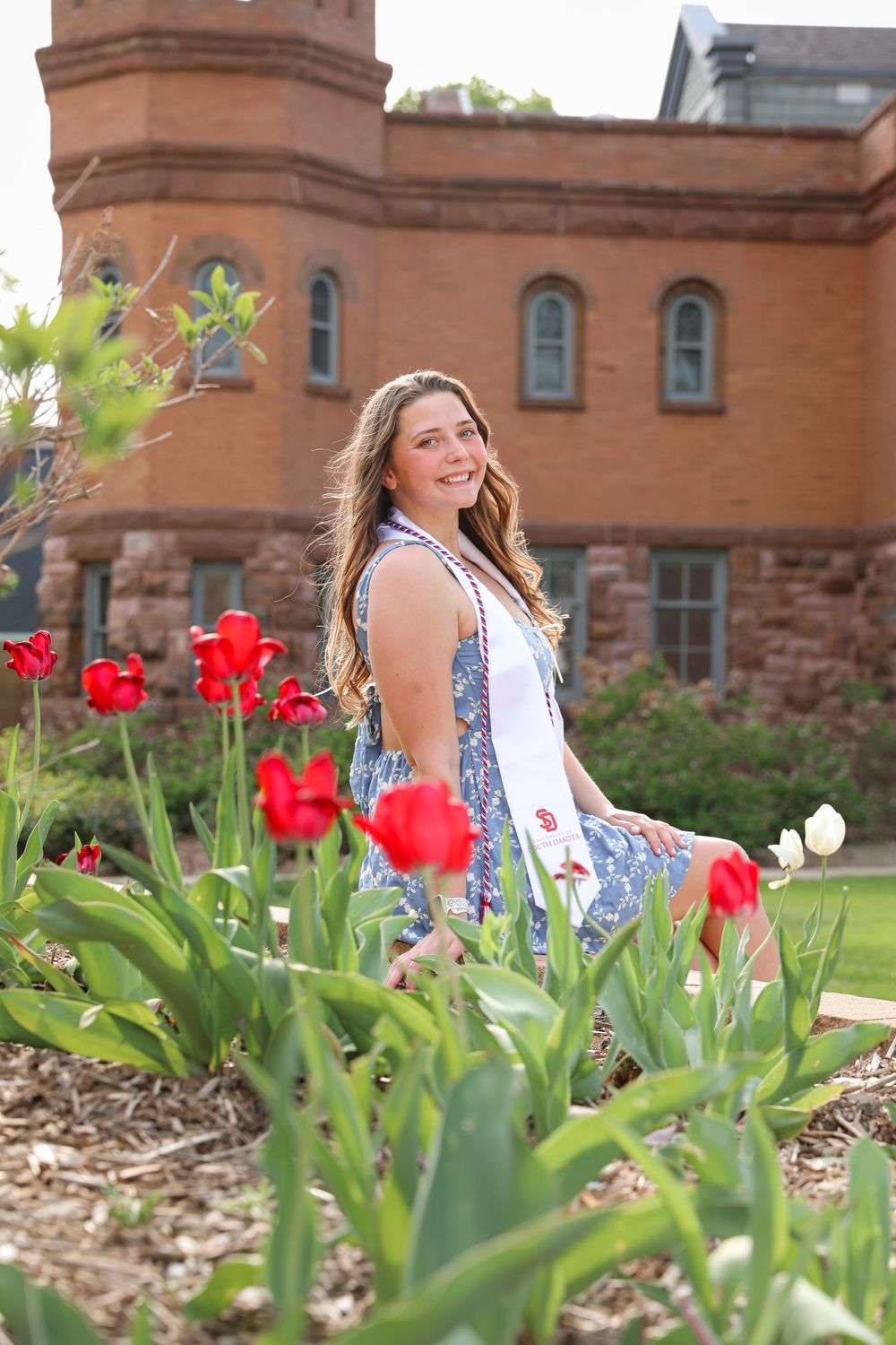 Young woman in floral dress sitting among red tulips in front of a historic red brick building.
