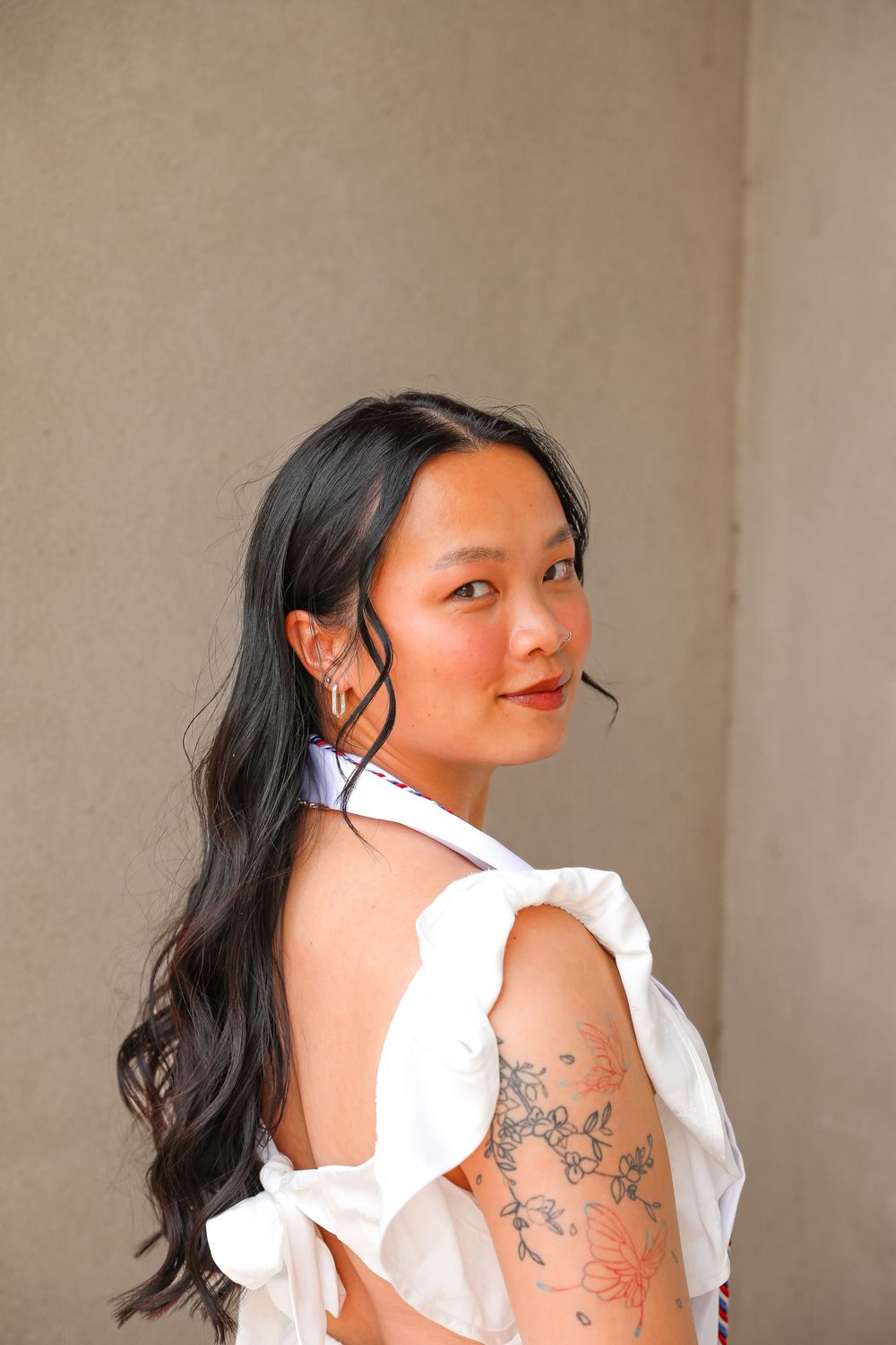 Young woman with dark hair in white backless top with floral tattoo on arm, looking over shoulder.