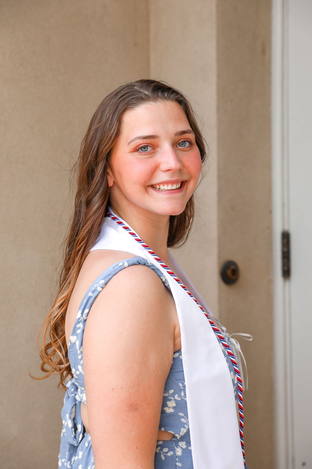 Smiling young woman wearing a white graduation stole with red and blue trim, posing outdoors in a floral dress.