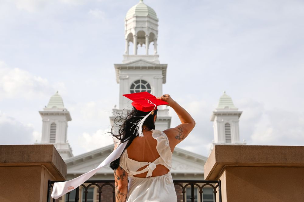 Graduate in white dress holds red cap before white building with towers on graduation day.
