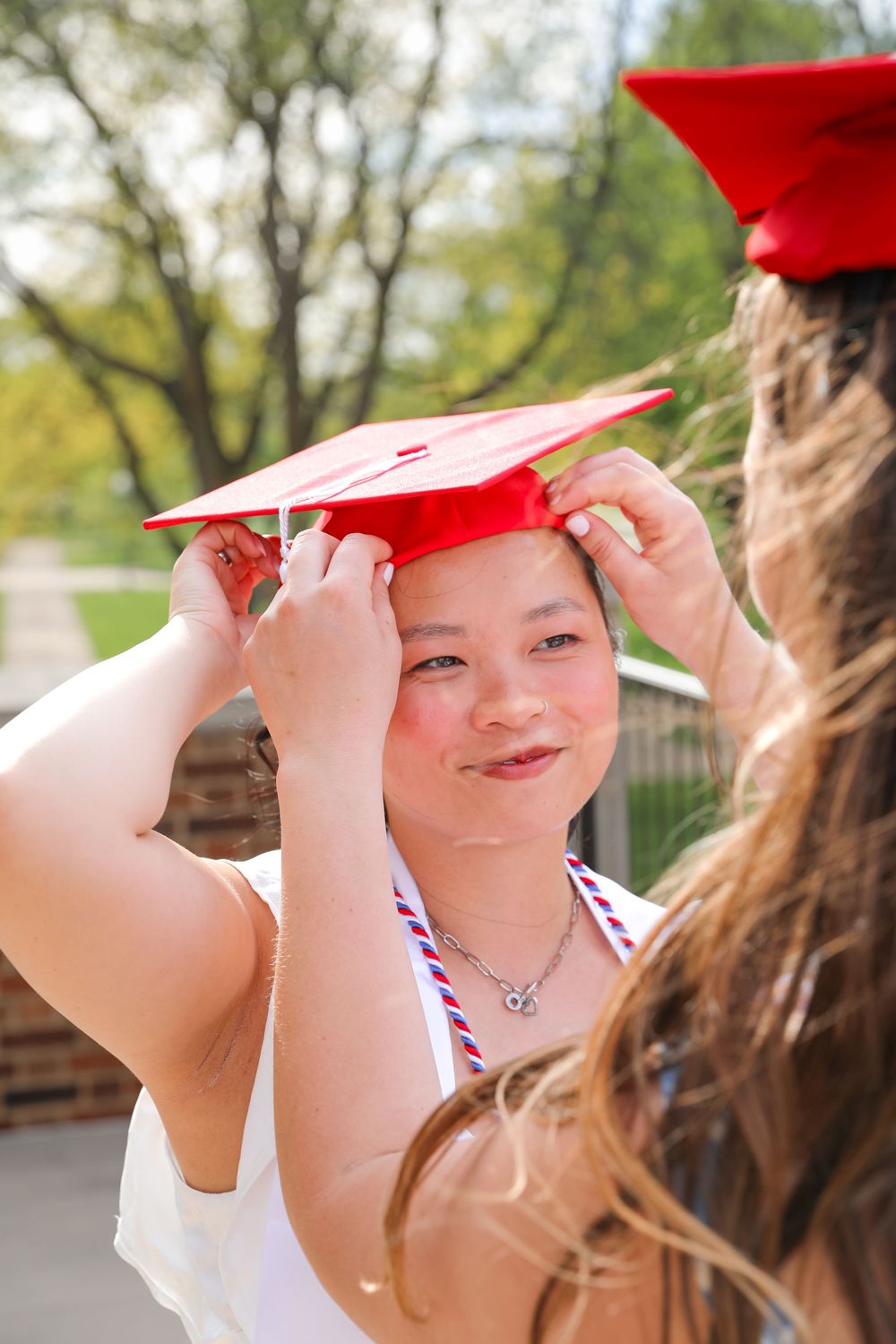 Young woman having red graduation cap adjusted by friend outdoors on a sunny day.