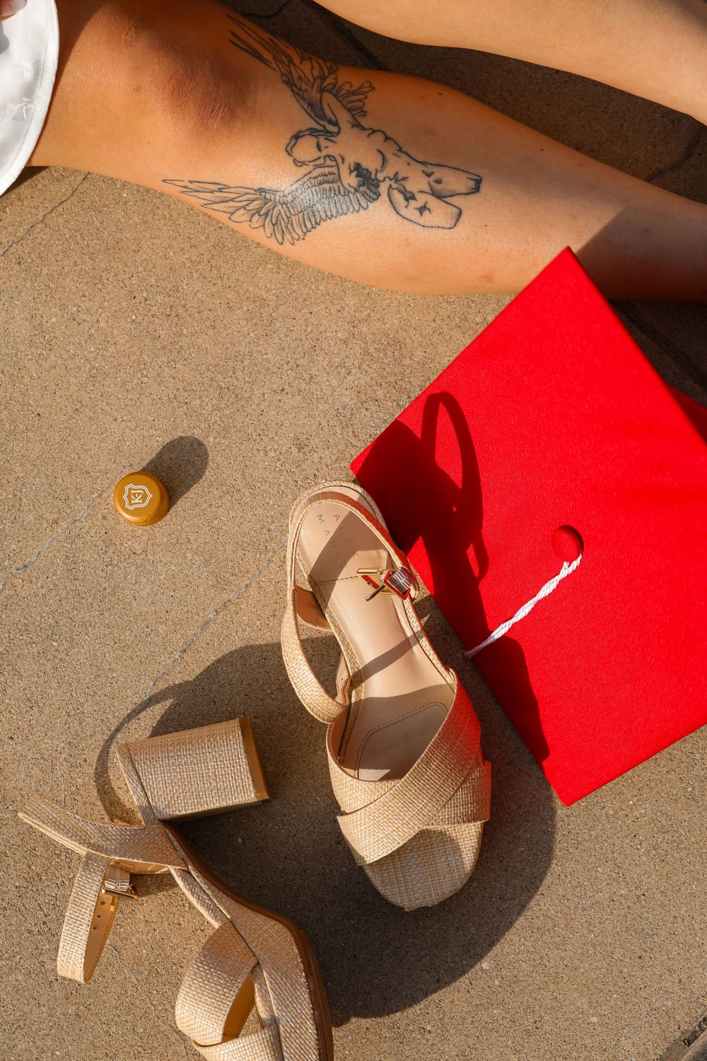 Beige platform sandals beside a red shopping bag, nail polish, and a tattooed arm on sandy ground.