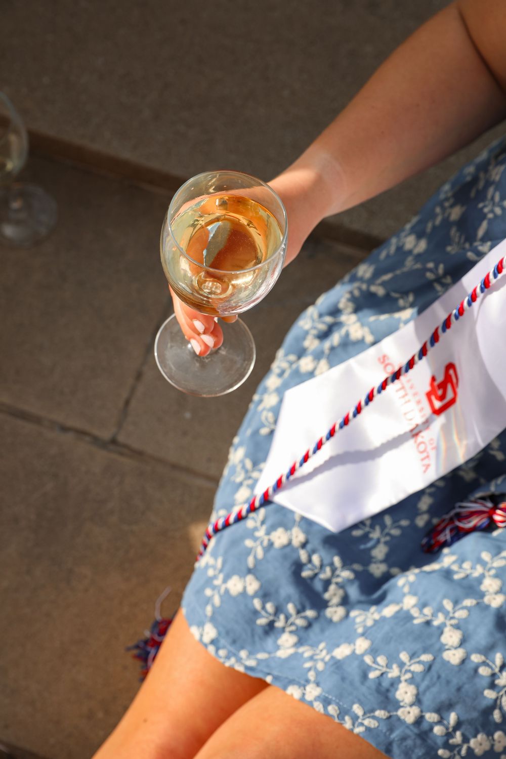 Woman in blue floral dress holding a glass of white wine outdoors in sunlight.