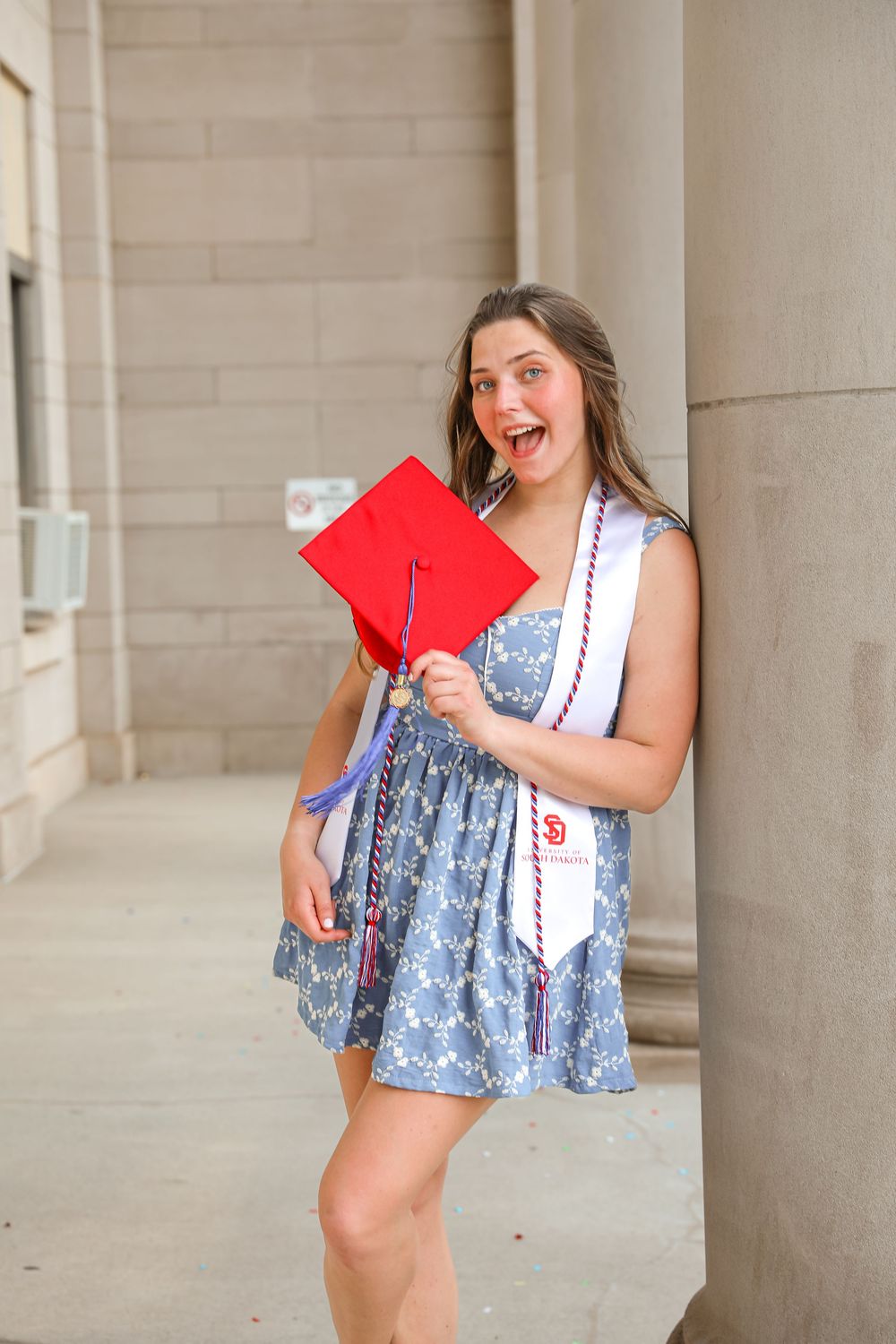 Excited young woman in floral dress holding red graduation cap and honor cords, leaning against stone column.