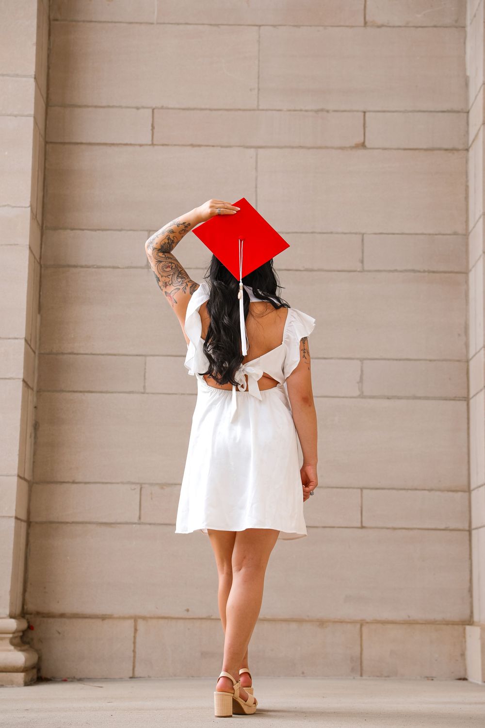Woman in white dress and red graduation cap posing against stone wall, showing tattooed arm.