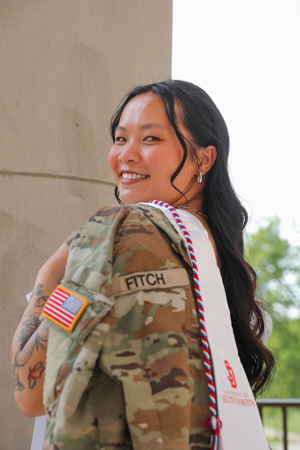 Smiling young woman in military camouflage uniform with Fitch name tape and US flag patch, wearing graduation sash.