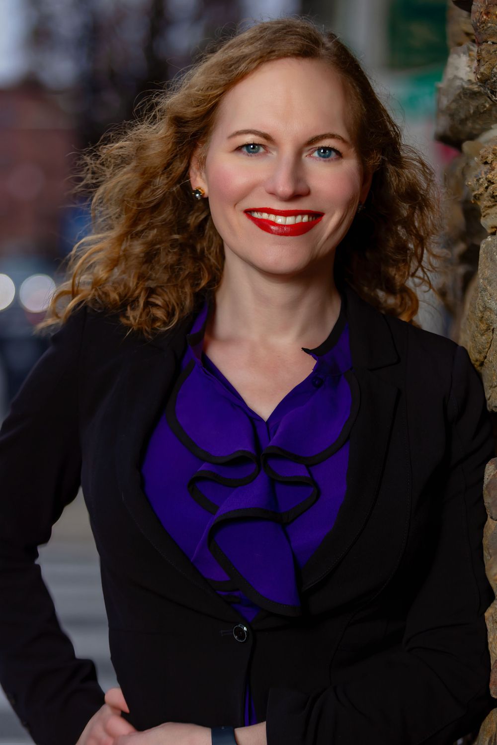 Professional portrait against dark background wearing black blazer and purple scarf.
