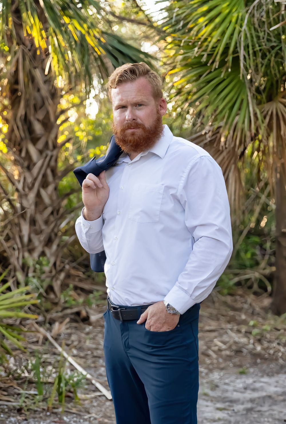 Man in white shirt and navy pants poses in tropical setting.