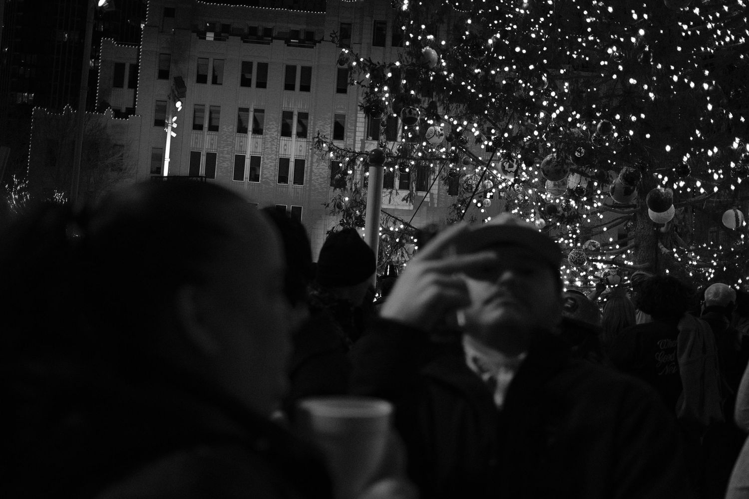 Black and white night photo of crowd gathered around a lit Christmas tree at an outdoor holiday event.