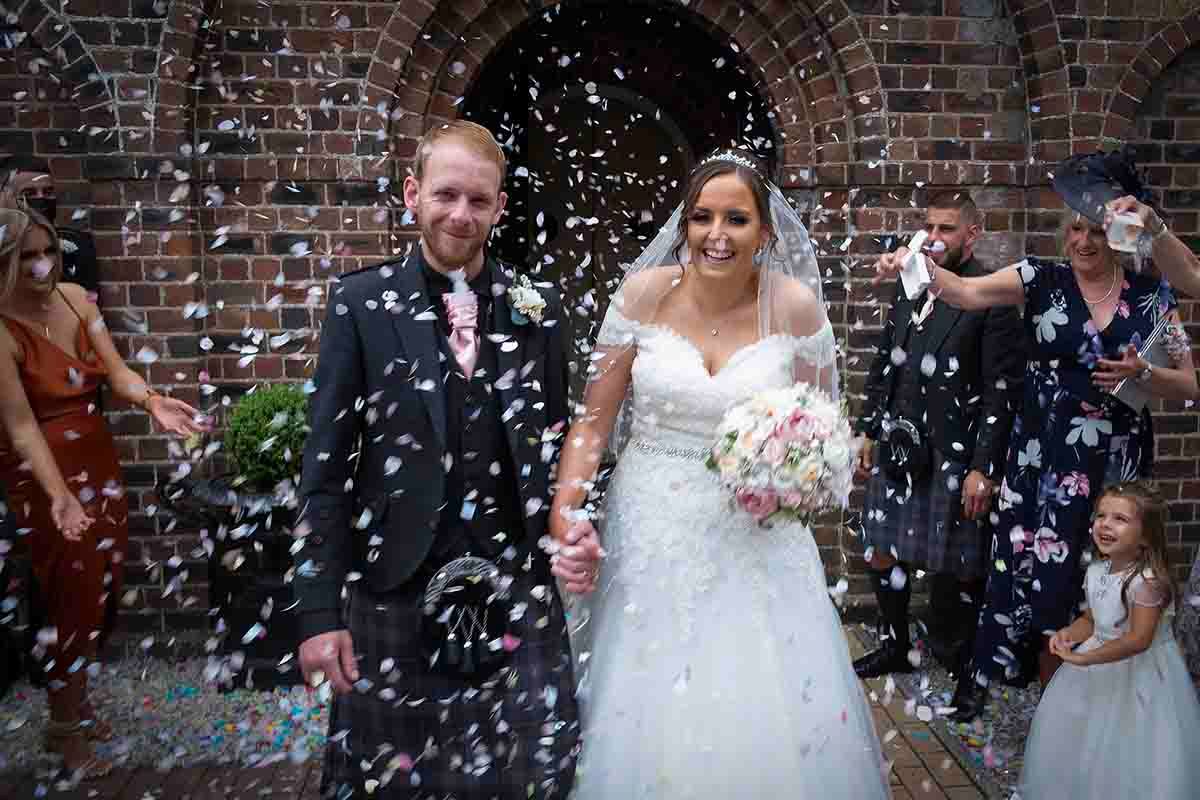 Bride and groom walk through confetti outside a brick church, surrounded by smiling guests and a flower girl at Gretna Green