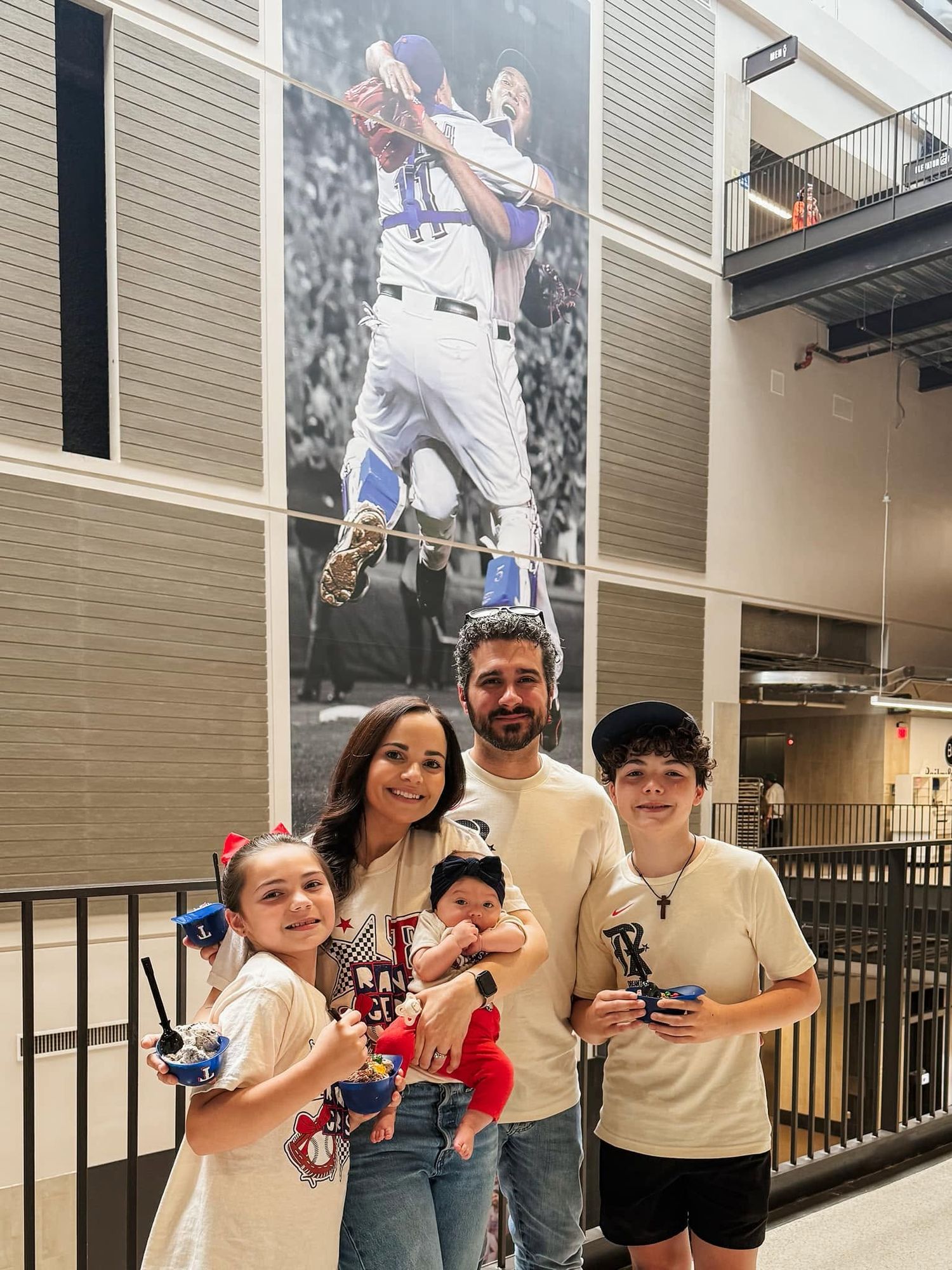 Family of five posing indoors at baseball stadium with large player mural on wall behind them.