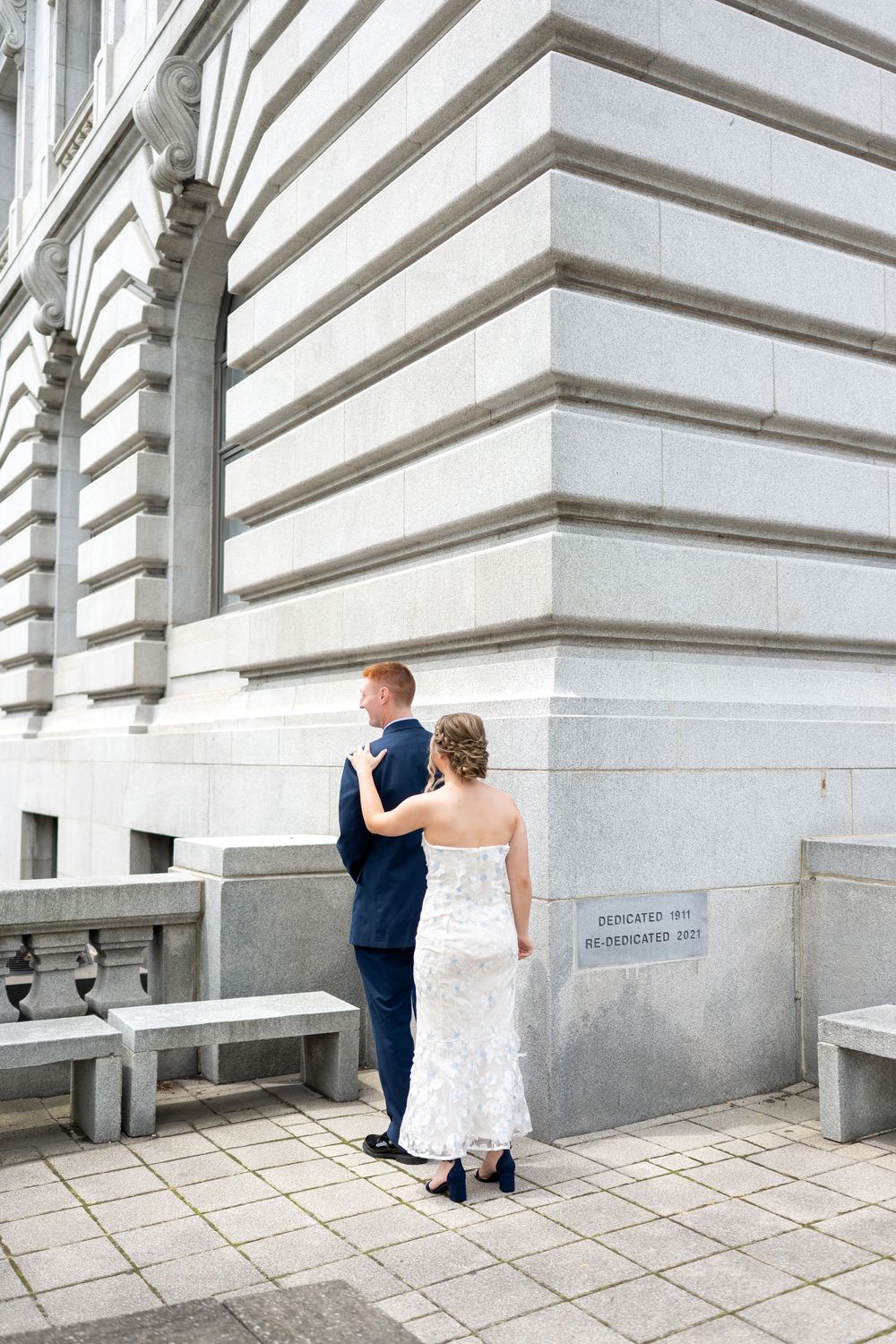 Bride in lace gown and groom in navy suit stand together at corner of grand stone building on wedding day.