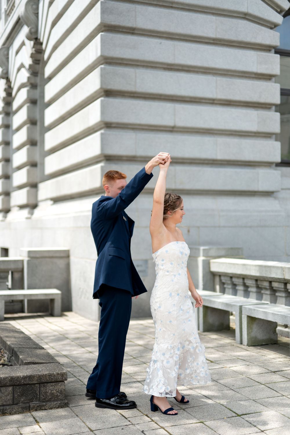 Groom in navy suit spinning bride in white lace wedding dress outdoors near elegant stone building.