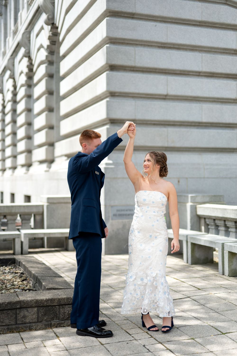 Bride and groom dancing outdoors near a stone building, groom in navy suit raising bride's hand as she smiles in white gown.
