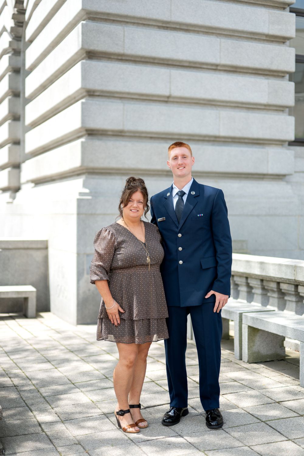 Young man in Air Force dress uniform poses with smiling woman in gray dress outside stone government building.