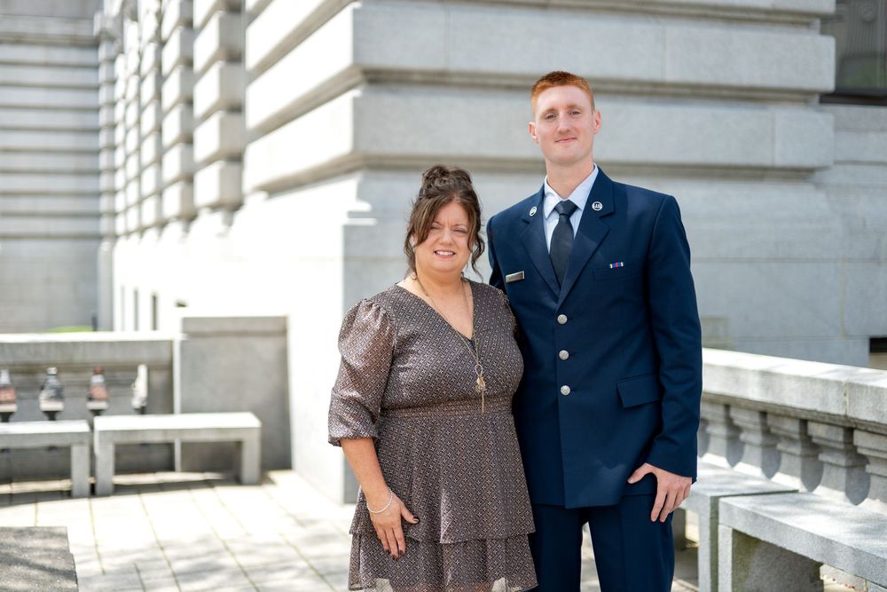 A woman in a floral dress and a young man in military dress uniform pose together outdoors near stone columns.