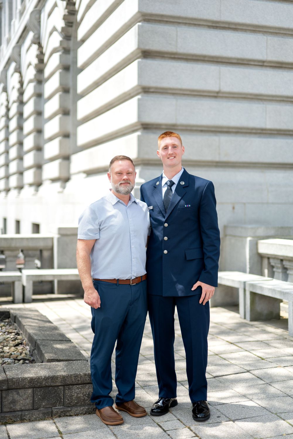 Two men pose outdoors near a government building, one in a navy military dress uniform and one in casual attire.