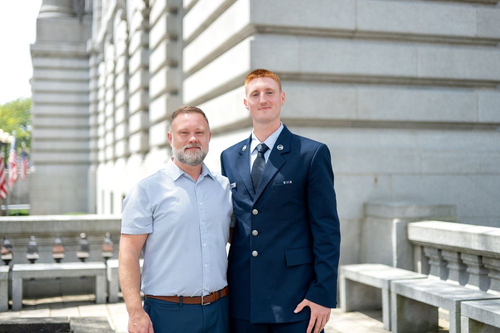 Two men posing outdoors near a government building, one in formal navy uniform with tie, the other in casual attire.