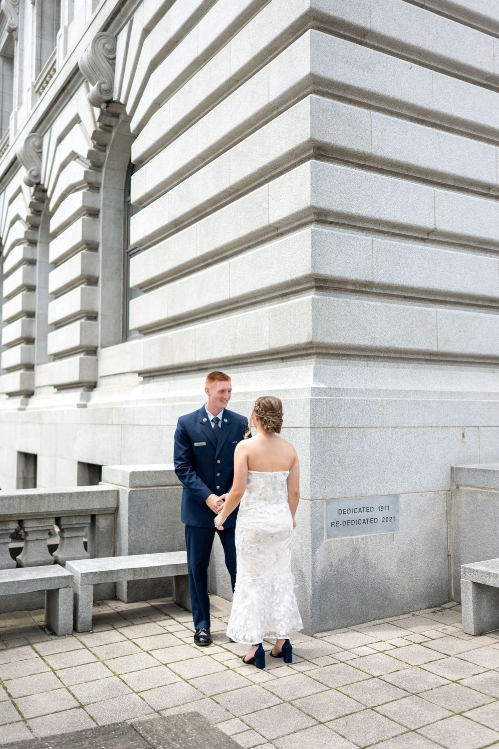 Military groom in blue uniform holds hands with bride in white lace dress outside a grand stone building.