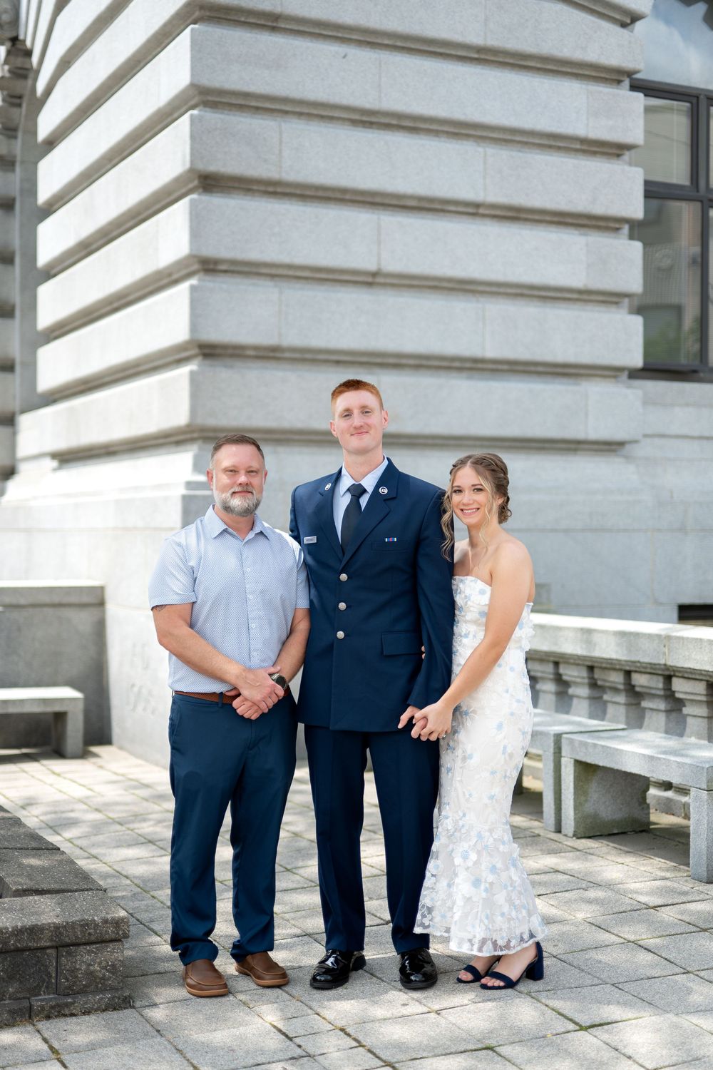 Air Force serviceman in dress uniform stands with man and woman in formal attire outside stone building.