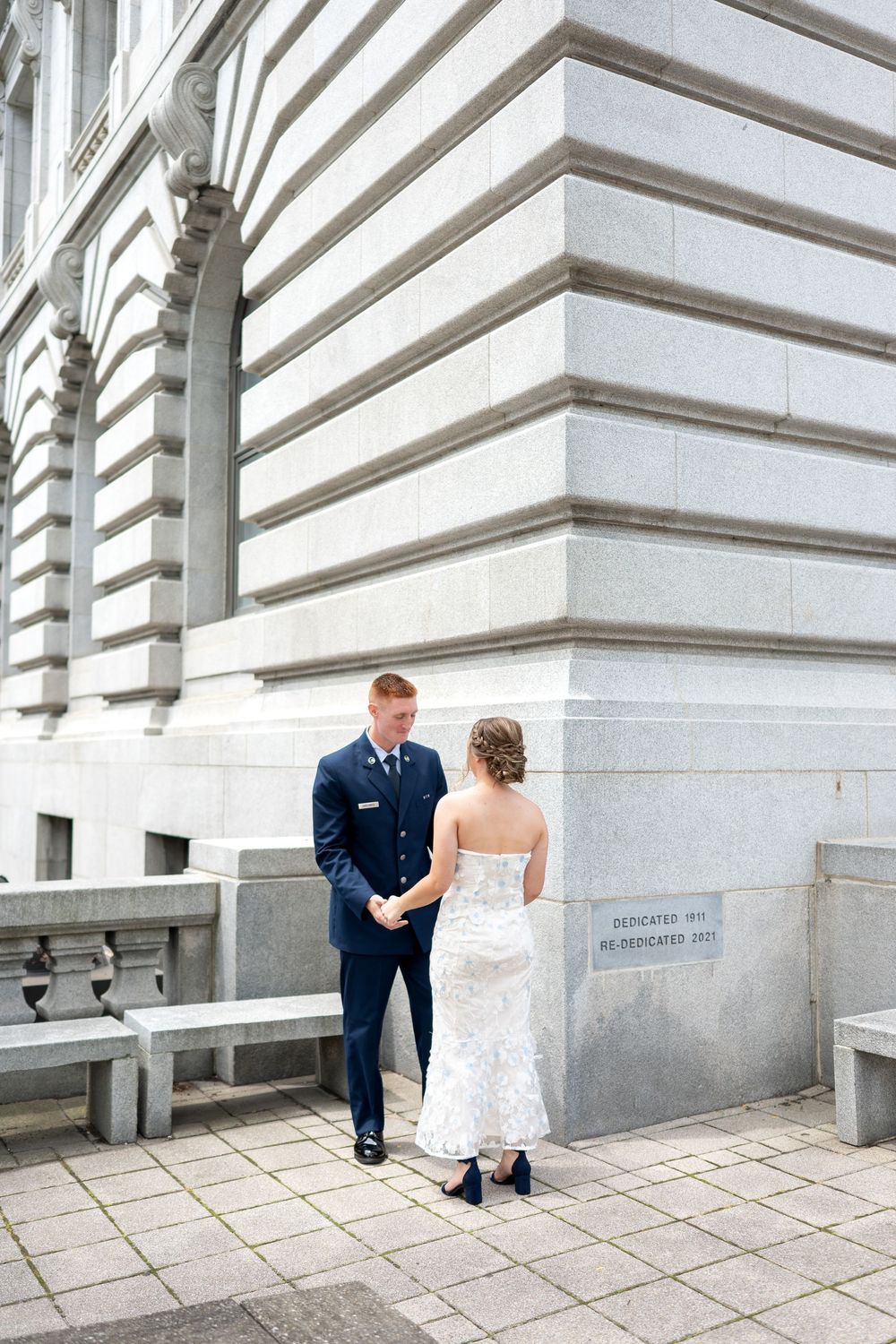 Military groom in navy uniform holds bride's hands in white lace gown outside grand stone building.
