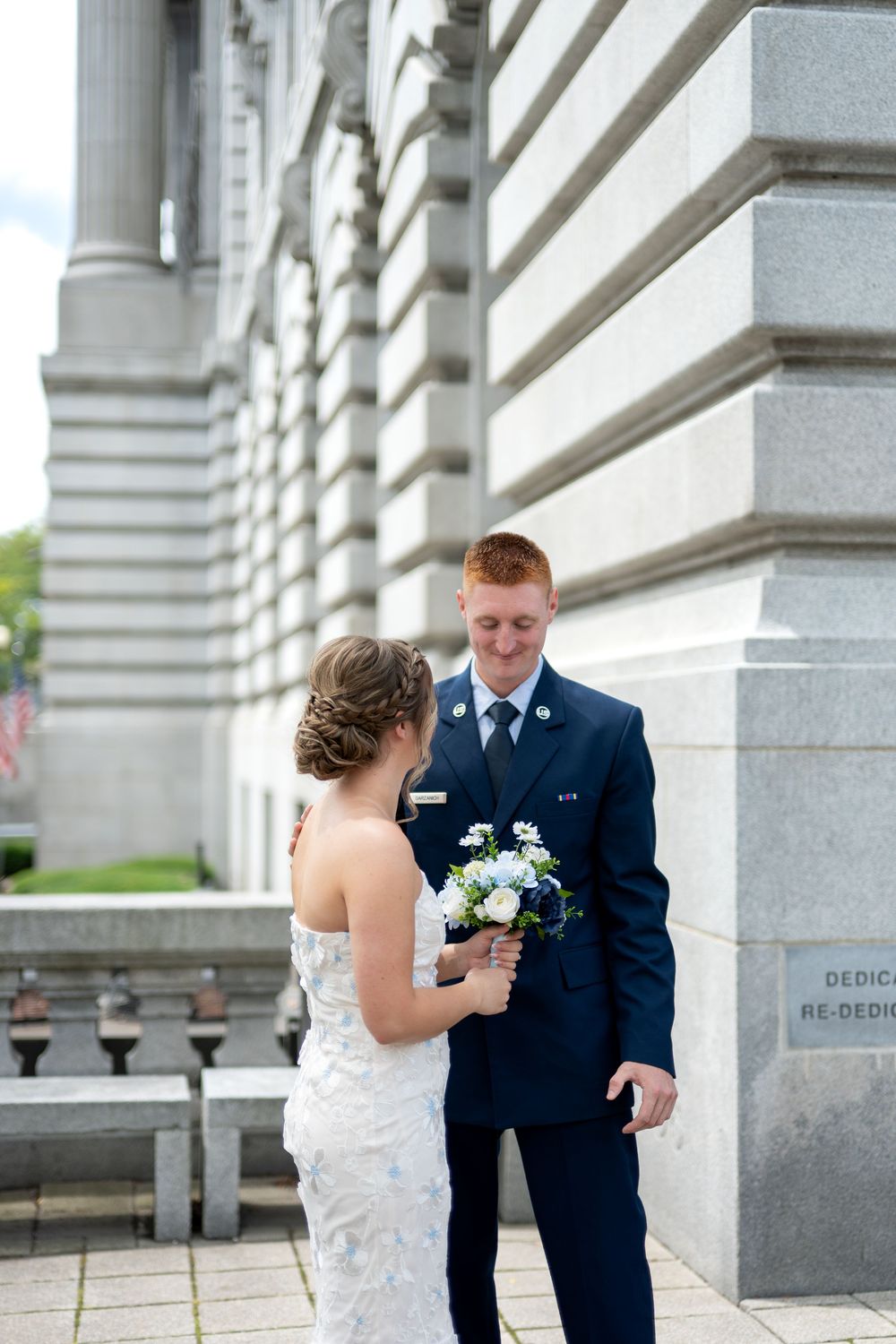 Military groom in navy suit smiles at bride in lace wedding dress holding white bouquet outside stone building.