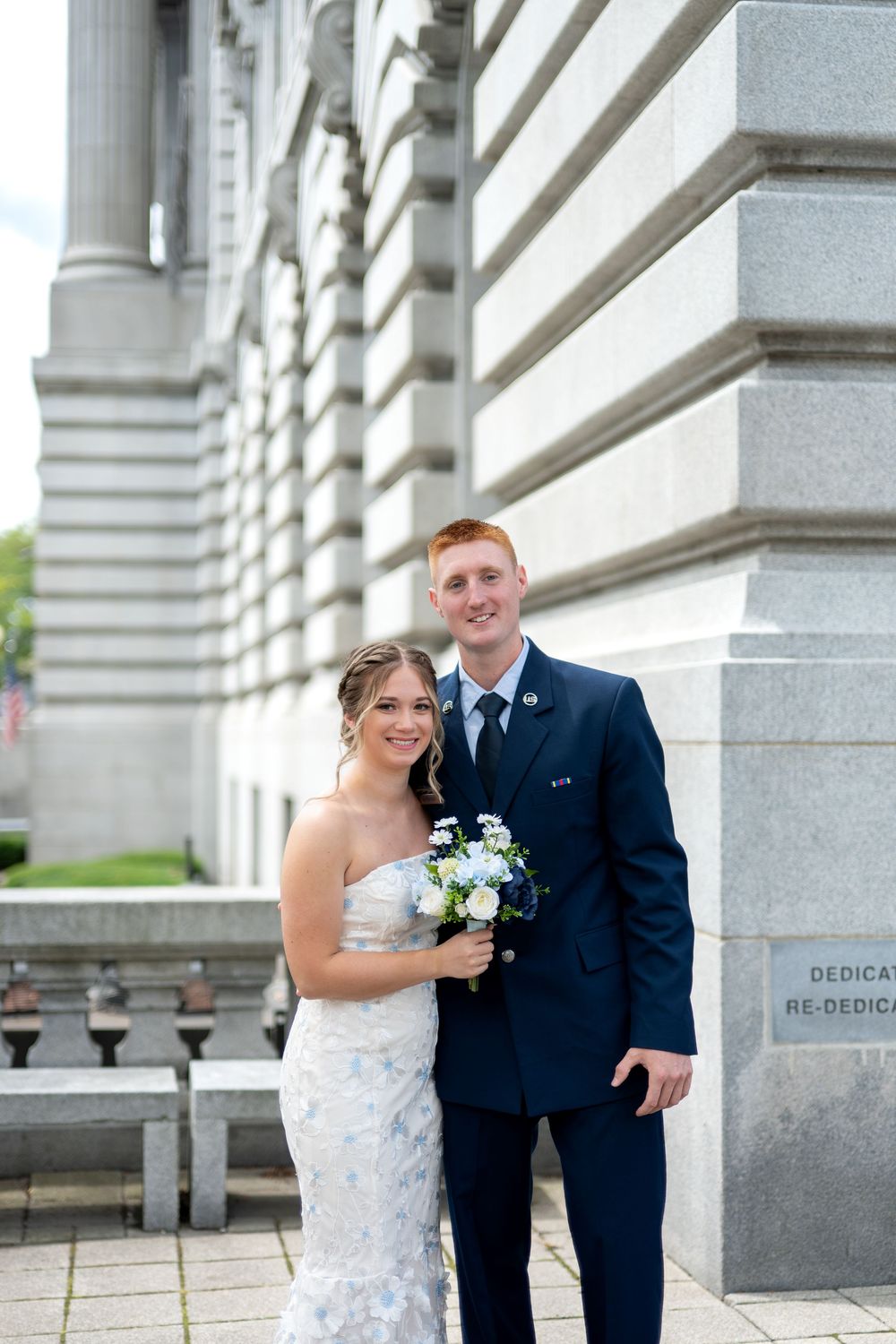 Bride and groom pose outside a grand stone building, she holds a white bouquet in a floral gown, he wears a navy suit.